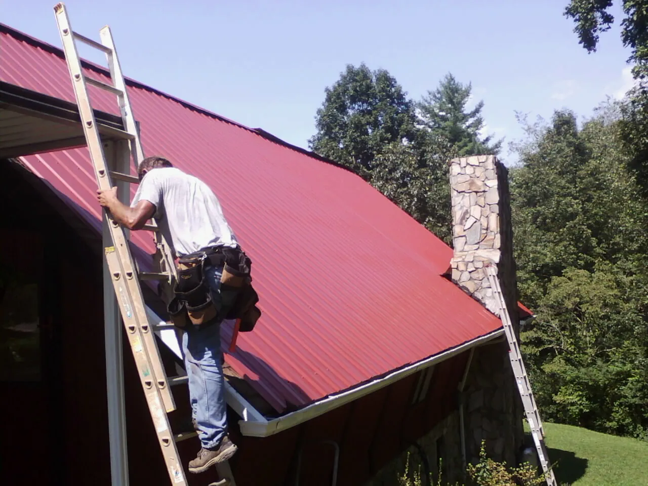 Roofer on a ladder beside a red metal roof and stone chimney — Soffit & Fascia Repair by Roofing Brothers in Bracey